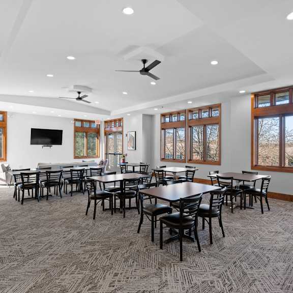 a clubhouse at new kent apartments in west chester, pa, featuring tables, chairs, a tv, carpet flooring, recessed lighting, two ceiling fans, and large windows offering beautiful views