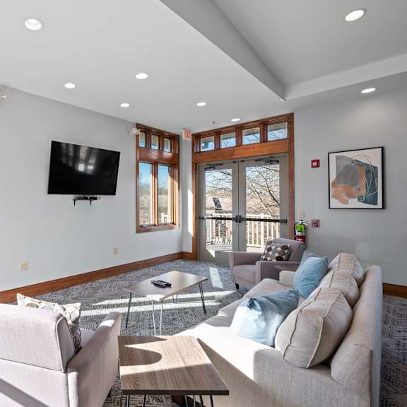 a community lounge at new kent apartments in west chester, pa, with cozy seating, coffee tables, a tv, and windows offering views of the outdoor green space