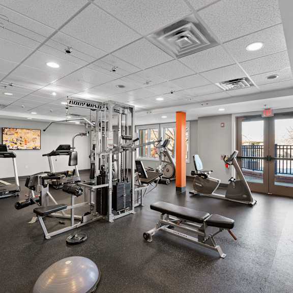 a spacious fitness center at new kent apartments in west chester, pa, featuring cardio and weight equipment, recessed lighting, large windows, a tv, and double doors opening to the outside