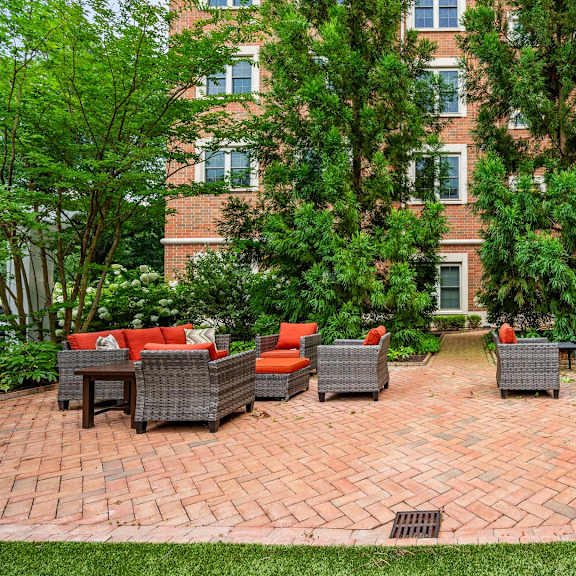 a patio with red cushions and grey wicker furniture is surrounded by trees and a brick building.