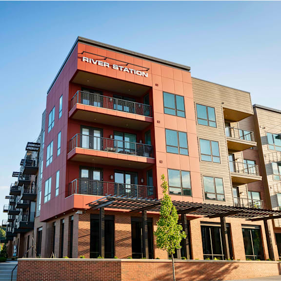 river station's four-story apartment building with balconies and a pergola with trees on the main level.