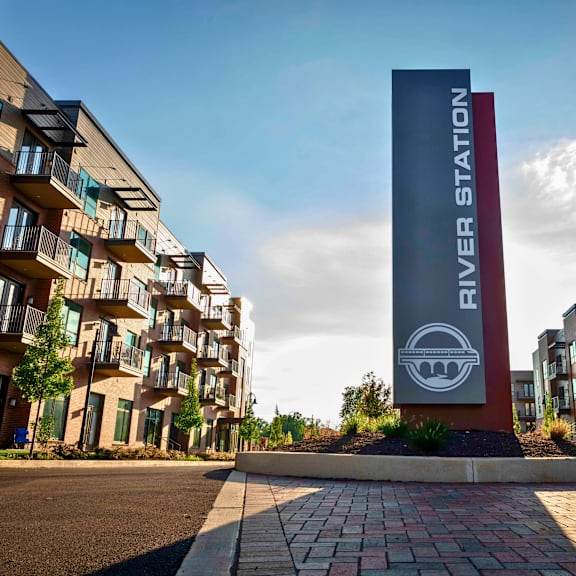 a river station sign in front of modern apartment buildings with balconies and trees lining a street.