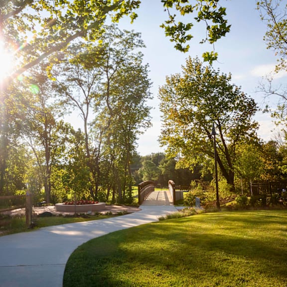 a beautiful trail with the sun shining through large trees and well-maintained grass leading to a bridge.