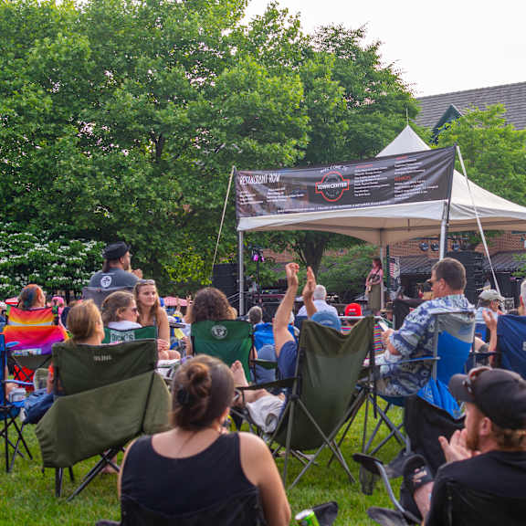a group of people are sitting in lawn chairs under a tent, watching a speaker.