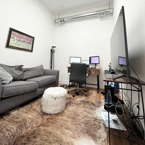 a room with a grey couch, a white ottoman, a desk with a computer and a monitor, and a picture on the wall.