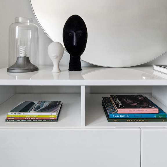 a round mirror on a white shelf with books and sculptures