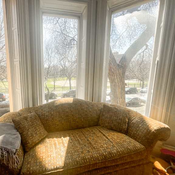a couch with a blanket on it is in front of a window with a view of a parking lot and trees.