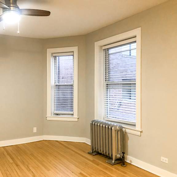 a room with a ceiling fan, two windows with blinds, and a radiator on the floor.
