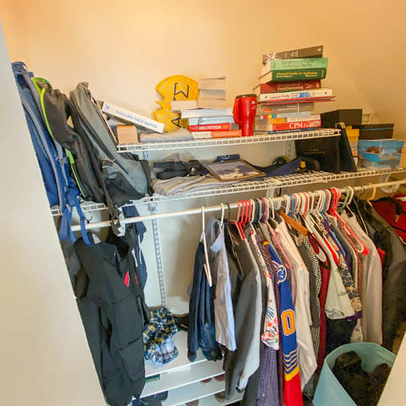 a cluttered closet with clothes and books on top.
