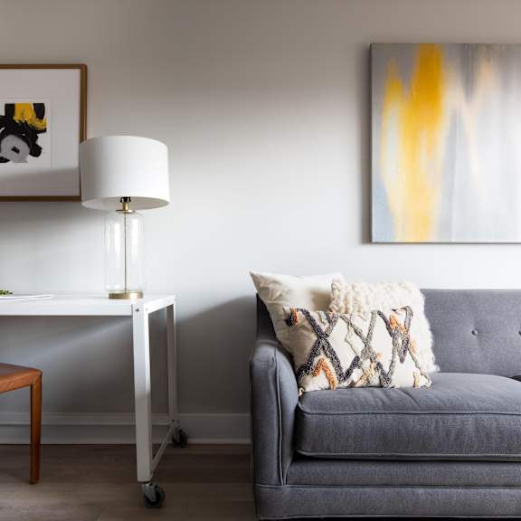 a living room with a grey sofa and a white desk with a lamp and a painting on