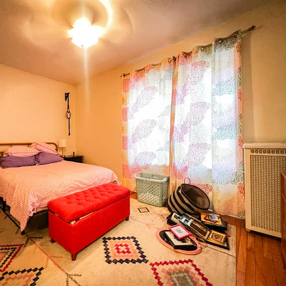 a bedroom with a bed, a red ottoman, and a rug with a geometric pattern.