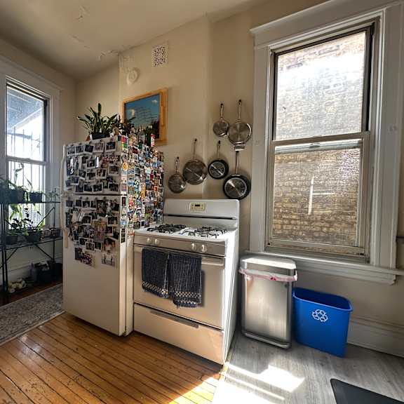a kitchen with a white refrigerator covered in magnets and notes, a stove with a pot on it, and a window with a view of a brick wall.