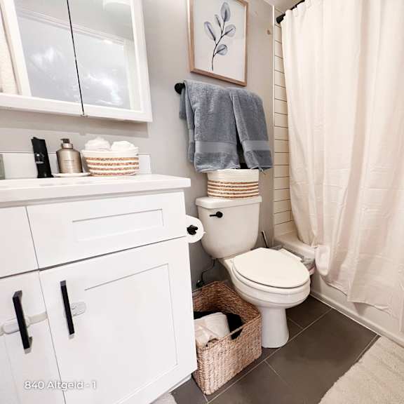 a white bathroom with a toilet, a shower curtain, and a basket of toiletries.