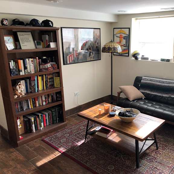a living room with a black leather couch, a wooden bookshelf filled with books, a coffee table with a bowl of fruit on it, and a rug on the floor.