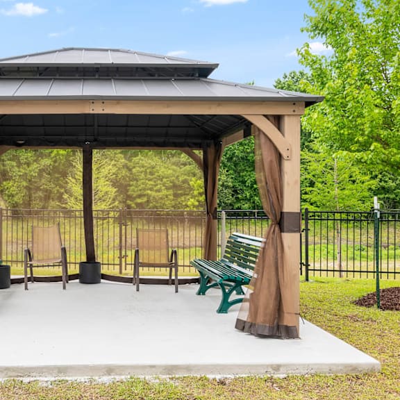 a gazebo with a brown canopy and green benches is surrounded by a black fence.