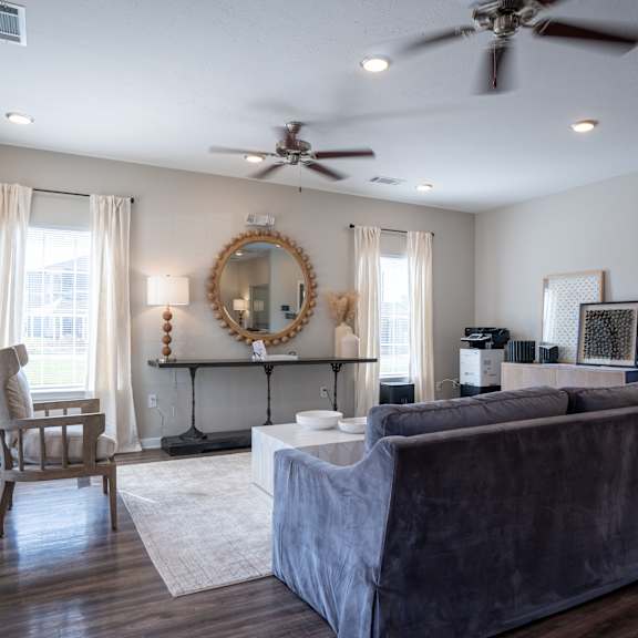 a living room with a grey couch, a chair, a mirror, and a ceiling fan.