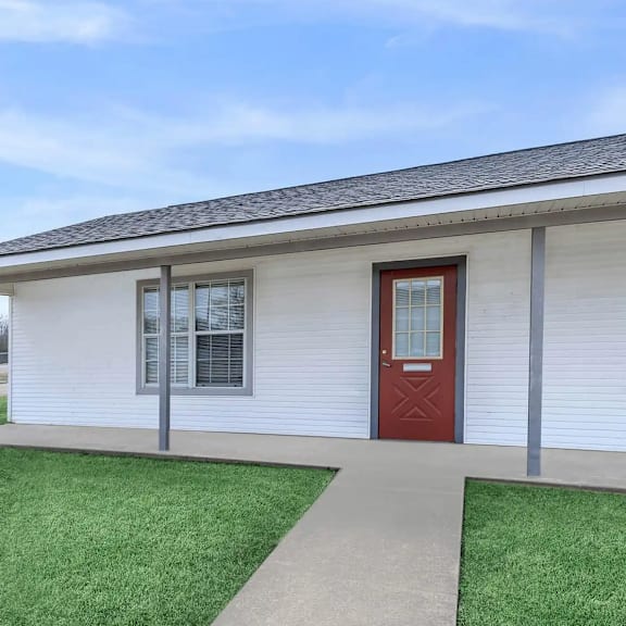 a small white house with a red door and a brown walkway leading to it.