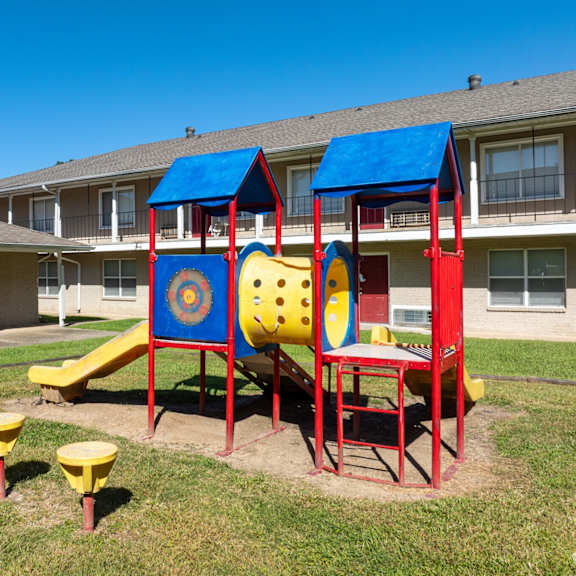 a playground with a slide, swings, and seesaw in front of a building.