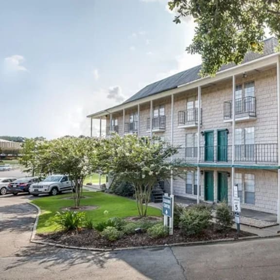 a building with a balcony and a green door is surrounded by trees and has a parking lot in front.