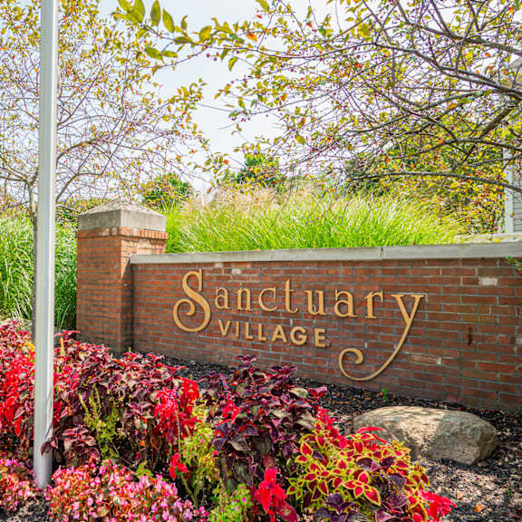 a sign for sanctuary village in front of a brick wall and flowers