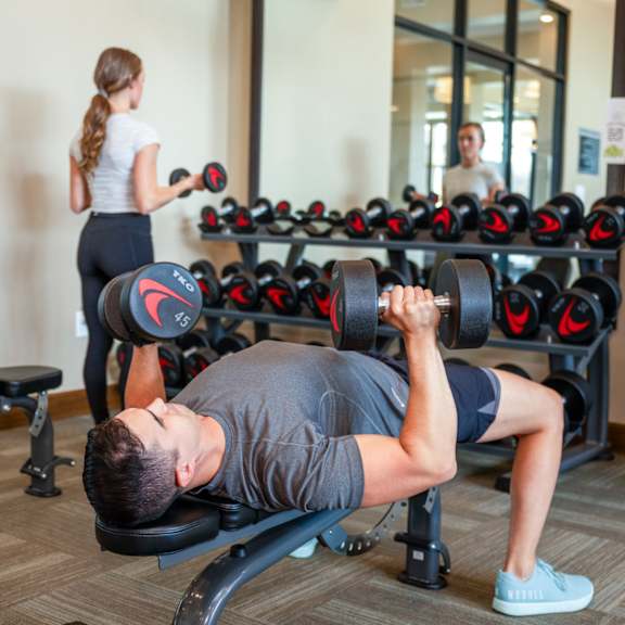 a man lifting weights in a gym with a woman in the background