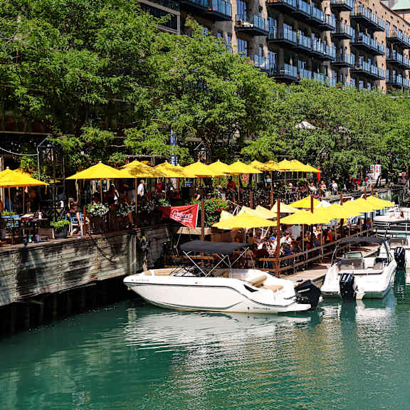 A row of boats are docked in a canal with yellow umbrellas and people sitting at tables in the background.