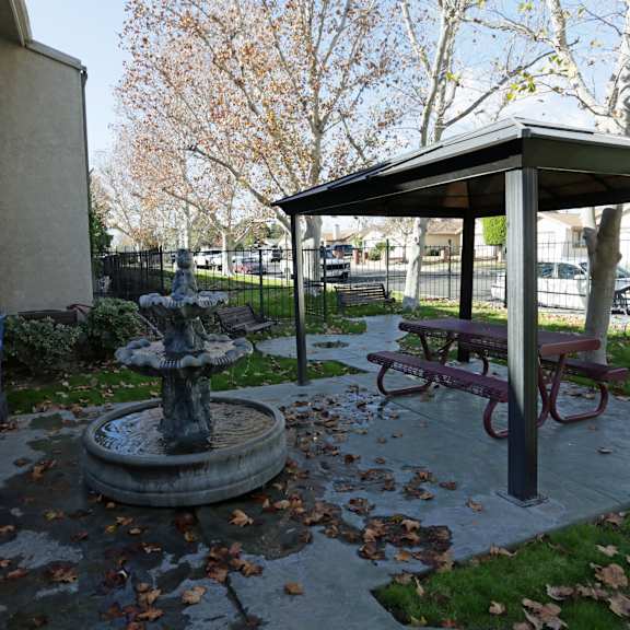 a small fountain in the middle of a park with a bench and a canopy nearby.