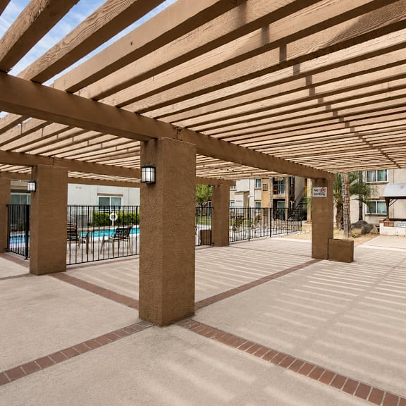 a patio with a wooden pergola and a pool in the background.