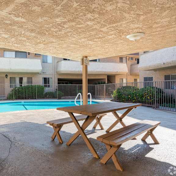a wooden picnic table is in the middle of a concrete courtyard with a pool and apartment buildings in the background.
