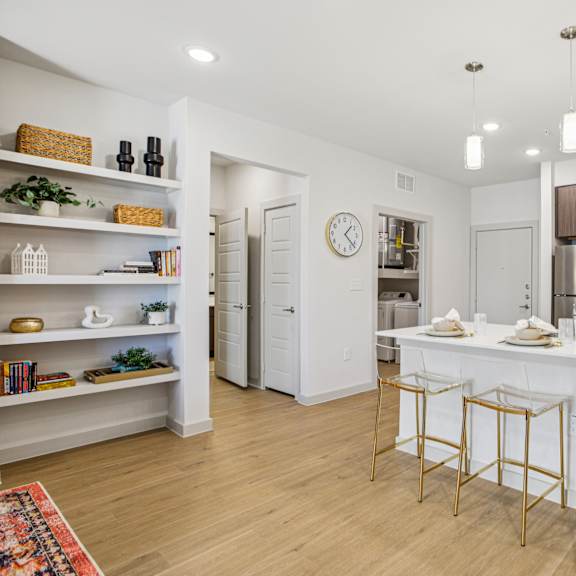 Thumbnail 16 of 37 A kitchen with white cabinets and a white island with gold bar stools at ThreeTwenty at the Vybe Apartments, Kyle, Texas