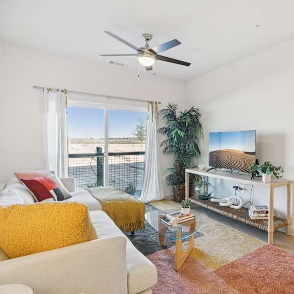 Thumbnail 6 of 23 A living room with a white couch, a yellow throw pillow, a TV, and a large window with a view of the outdoors. at ThreeTwenty at the Vybe Apartments, Texas