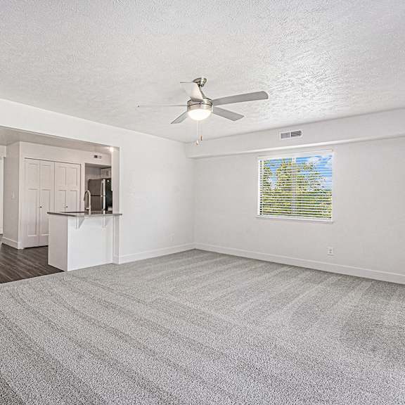Thumbnail 6 of 14 An enlarged living room with a ceiling fan and carpeted floor at Byron Lakes Apartments, Michigan, 49315