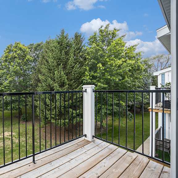 Thumbnail 5 of 19 A balcony with a black railing overlooks a grassy area with trees at Byron Lakes Apartments in Byron Center, MI