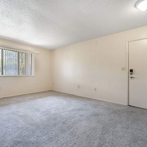 Thumbnail 4 of 14 A garden style living room with a carpeted floor, a window with blinds, and a door at Concord Place Apartments, Kalamazoo, MI, 49009