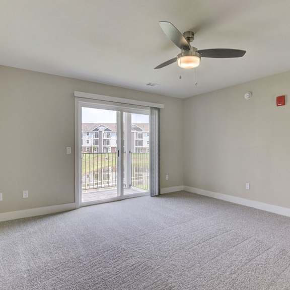 Thumbnail 5 of 14 A living room with a ceiling fan and carpeted floor leading to a private balcony at Copper Creek Apartment Homes, Maize, Kansas