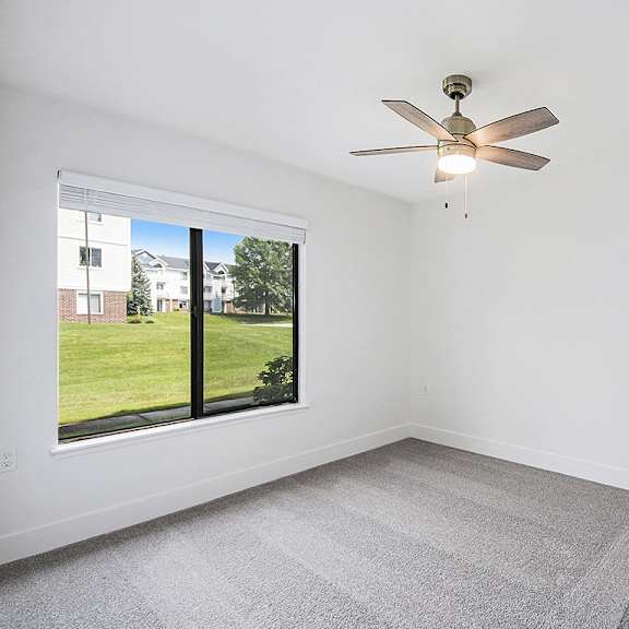 Thumbnail 12 of 15 A bedroom with a ceiling fan and a window overlooking a green lawn at The Crossings Apartments in Grand Rapids, MI