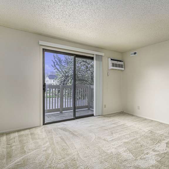 Thumbnail 3 of 8 A living room with a carpeted floor and a sliding glass door leading to a balcony at Madeira Apartments, Michigan