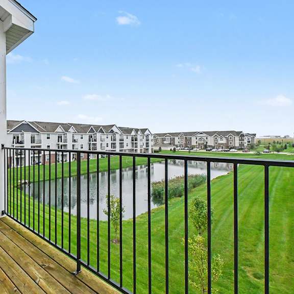 Thumbnail 5 of 16 A balcony with a black railing overlooks a grassy area with a large pond at The Reserve at The Reserve at Destination Pointe in Grimes, IA 50111