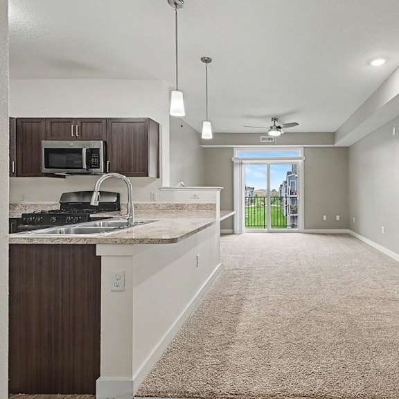 Thumbnail 6 of 16 A kitchen with a breakfast bar and a living room with a sliding glass door leading to a balcony at The Reserve at The Reserve at Destination Pointe in Grimes, IA 50111