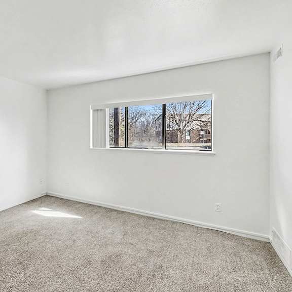 Thumbnail 3 of 11 A living room with a carpeted floor and a window showing a view of trees outside at Trappers Cove Apartments, Lansing