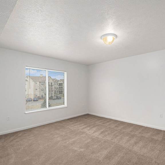 Thumbnail 14 of 19 A large bedroom with a carpeted floor and a window showing a view of buildings outside at Trillium Pointe Apartment Homes, Michigan, 49201
