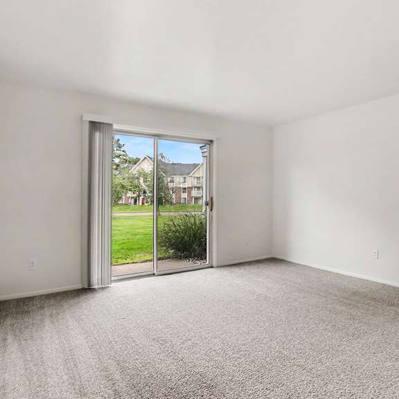 Thumbnail 3 of 11 A living room with a carpeted floor and a sliding glass door leading to a patio and green lawn at Walnut Trail Apartments, Michigan
