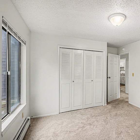 Thumbnail 15 of 15 A bedroom with a window, closet doors, and a carpeted floor at Waverly Park Apartments, Lansing, Michigan