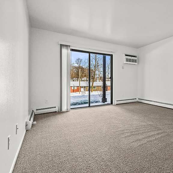 Thumbnail 3 of 14 A living room with a carpeted floor and a sliding glass door leading to a patio at Wood Creek Apartments, Kenosha