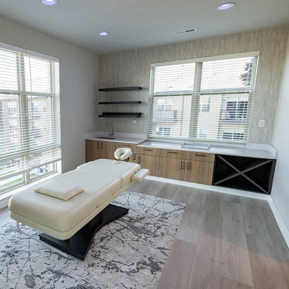 a room with a white massage table, wall shelving, and windows.