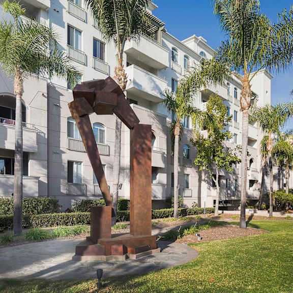 a sculpture of a man holding a woman is in the foreground of a grassy area in front of a building with palm trees. at la jolla crossroads apartments, california