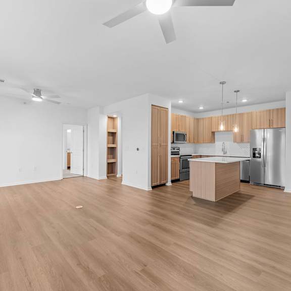Thumbnail 1 of 6 A spacious room with wooden flooring and a kitchen area in the background.