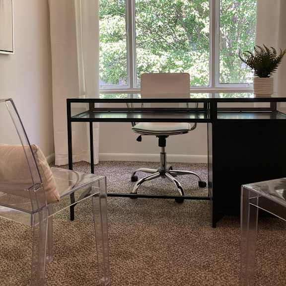 Thumbnail 6 of 8 A black desk with a glass top and a white chair in front of a window with trees visible outside.