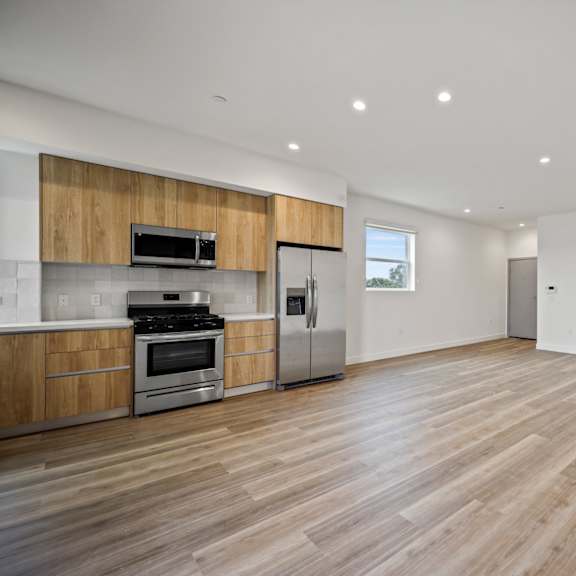 an empty living room with wood flooring and a kitchen with stainless steel appliances