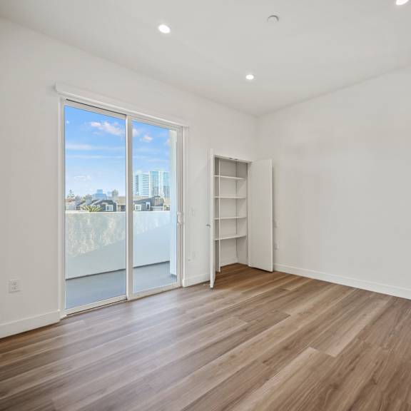 a room with a sliding glass door leading to a balcony, wooden flooring, and a white wall with a shelf.